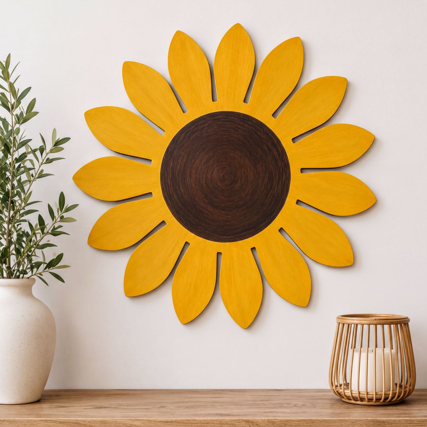 a large, vibrant yellow sunflower-shaped wall decoration hanging on a white wall. Next to the sunflower is a white vase with greenery, and a small woven basket with a candle inside.